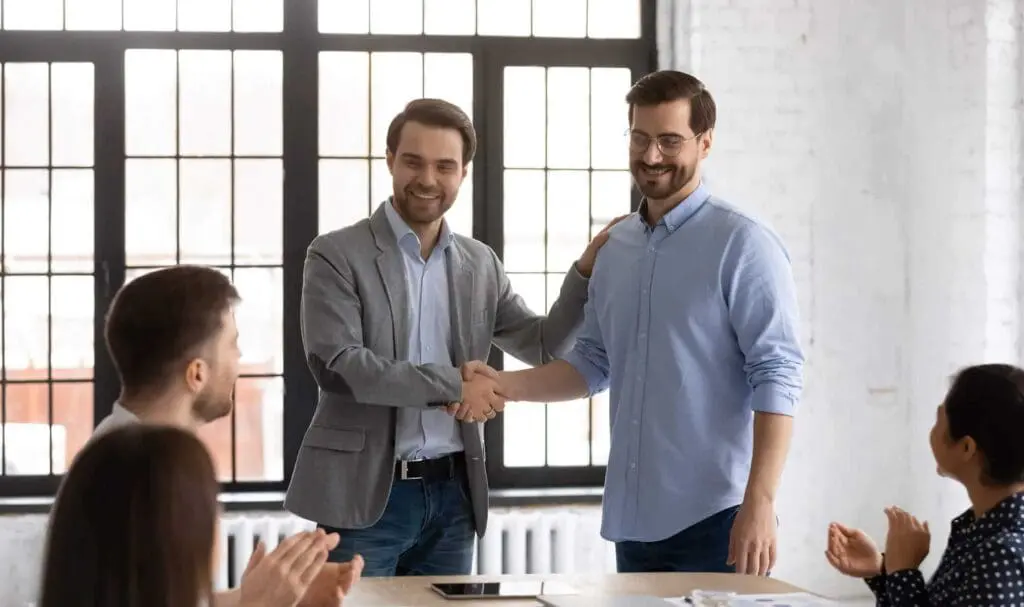 Deux hommes se tiennent debout et se serrent la main en souriant, tandis que trois collègues assis à une table les applaudissent dans un bureau lumineux avec de grandes fenêtres en arrière-plan.