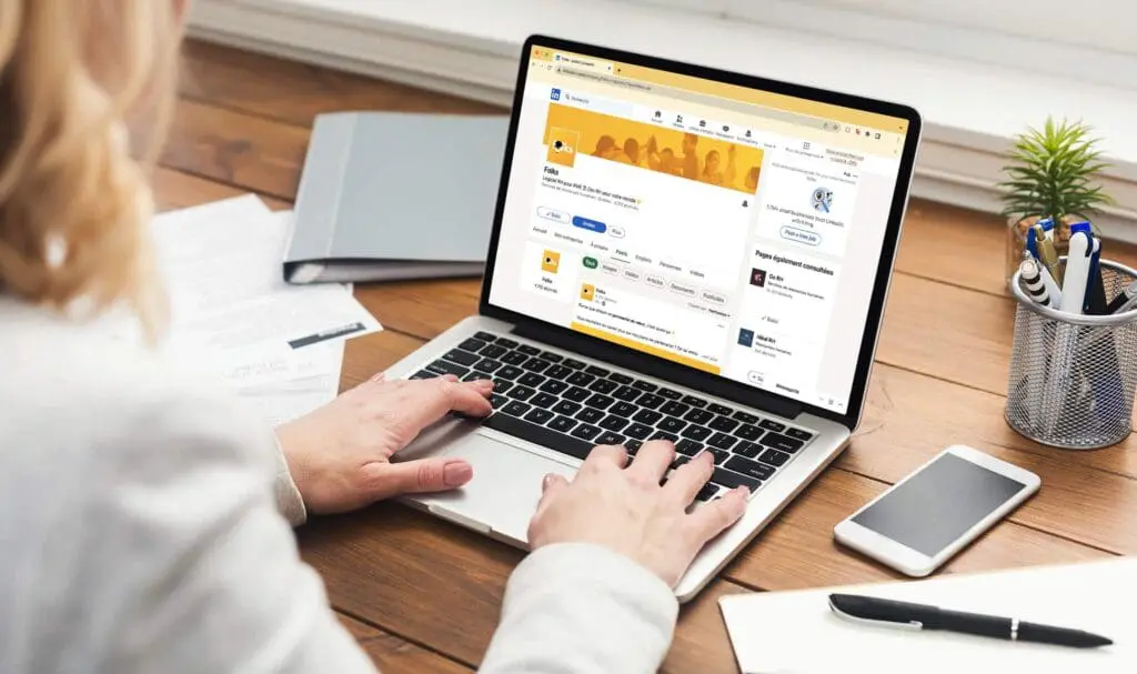 Une personne tape du texte sur un ordinateur portable affichant son profil LinkedIn. L'ordinateur est posé sur un bureau en bois, avec des papiers, un carnet fermé, un smartphone, un stylo et une petite plante en pot à proximité.