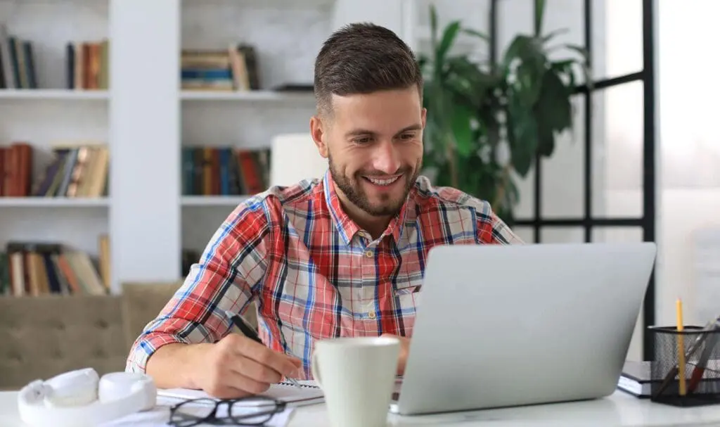 Un homme en chemise à carreaux sourit tout en travaillant sur un ordinateur portable à un bureau avec un cahier, un stylo, une tasse à café et des verres, dans une pièce lumineuse avec des étagères de livres et une grande plante en arrière-plan.