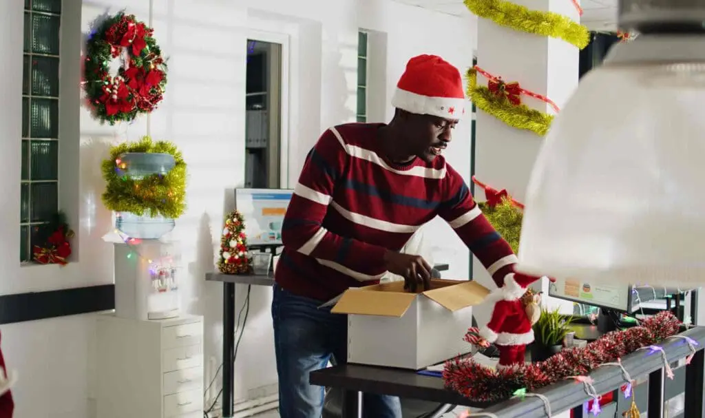 Un homme portant un bonnet de Père Noël et un pull rayé ouvre une boîte en carton sur un bureau décoré, entouré de décorations de Noël, de guirlandes et de décorations festives dans un cadre de bureau.