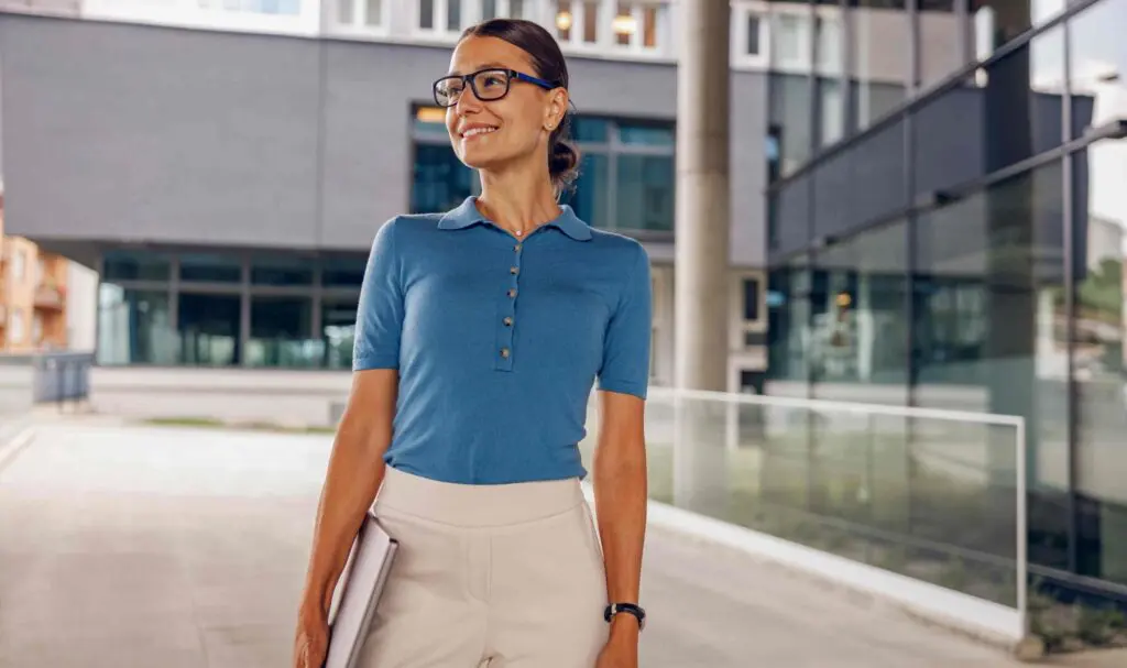 Une femme portant des lunettes, un polo bleu à manches courtes et un pantalon beige, se tient à l'extérieur devant un bâtiment moderne en verre, tenant un ordinateur portable fermé et souriant.