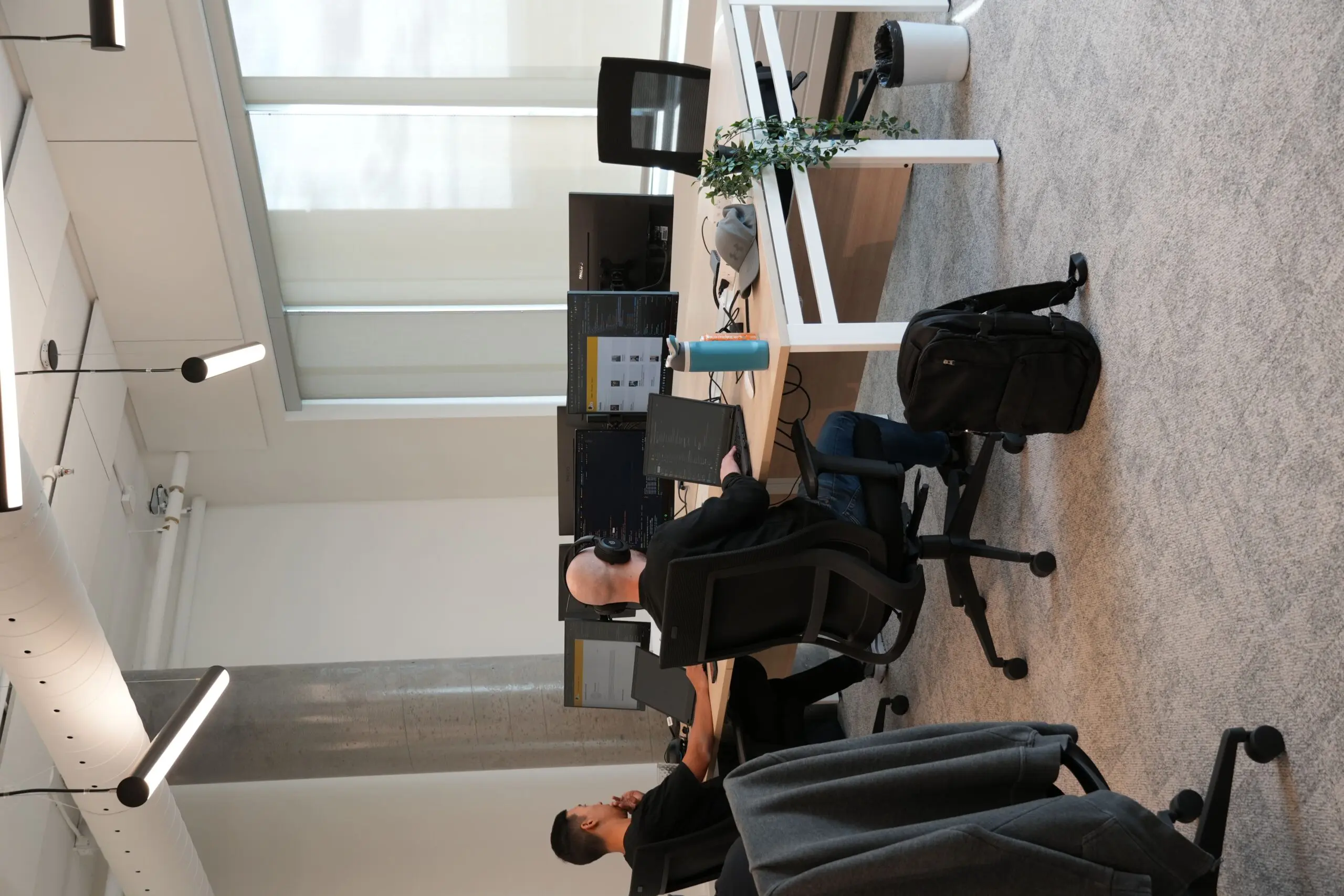 Two people work at desks with multiple monitors in a modern office. One person wears headphones, a backpack is on the floor, and a water bottle and plant are on the desk. Large windows let in natural light.