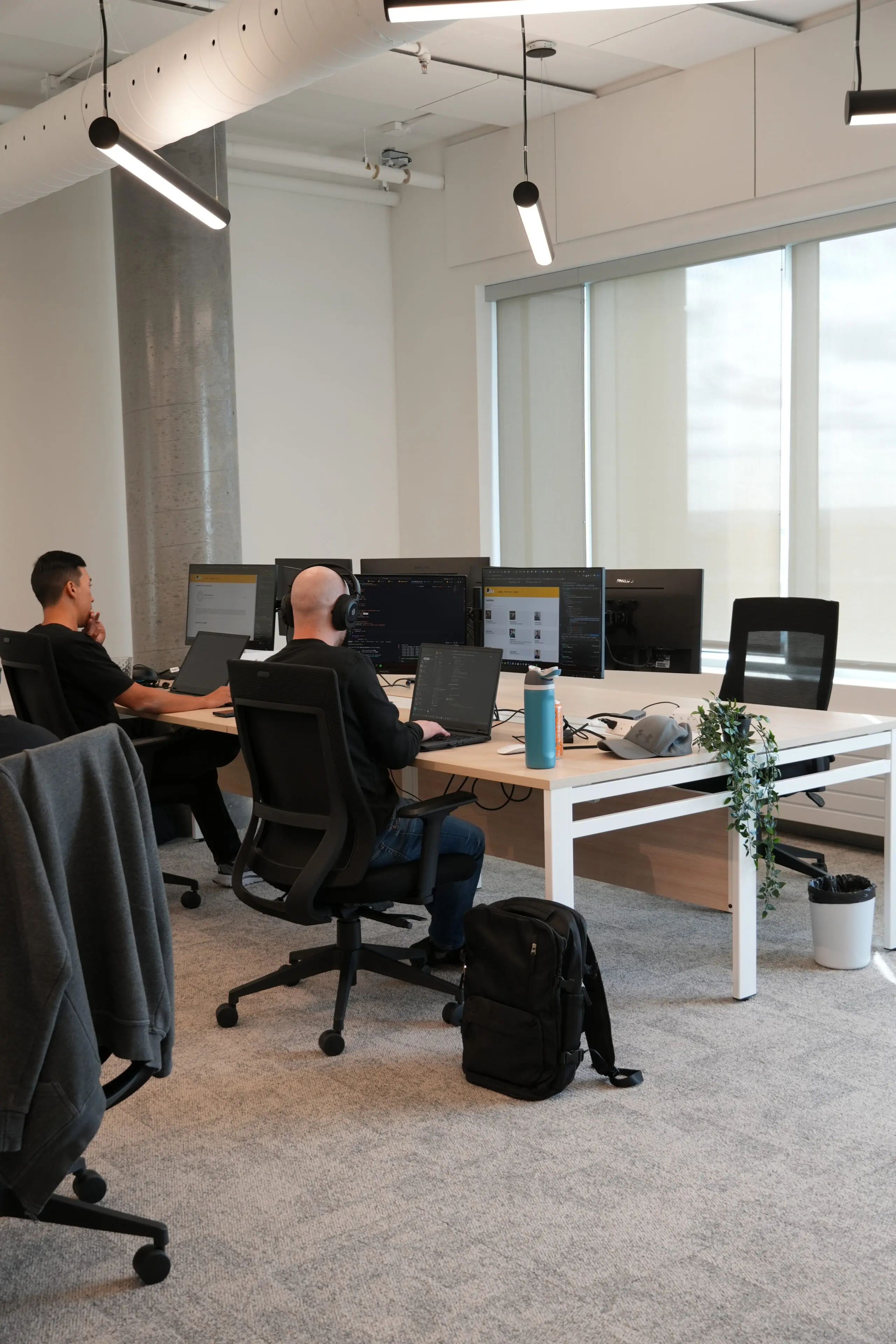Two people work at desks with multiple monitors in a modern office. One person wears headphones, a backpack is on the floor, and a water bottle and plant are on the desk. Large windows let in natural light.