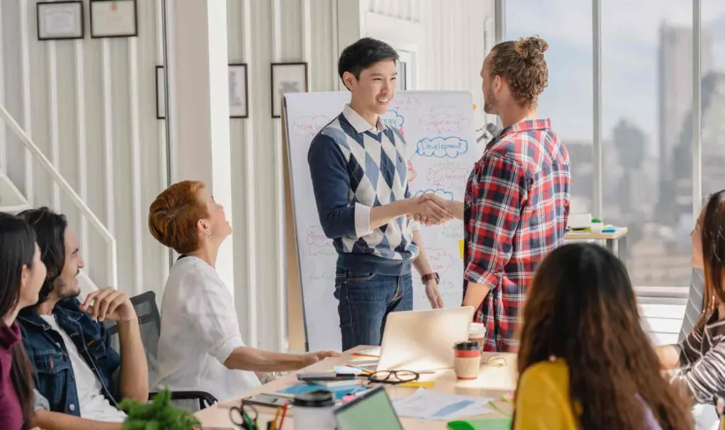 Un groupe de collègues est assis autour d'une table dans un bureau lumineux, tandis que deux personnes se serrent la main devant un tableau blanc couvert de notes et de diagrammes. Ordinateurs portables, carnets et tasses à café sont posés sur la table.