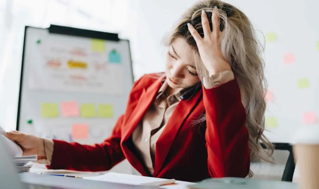 Une femme vêtue d'un blazer rouge est assise à un bureau, l'air stressée, la main dans les cheveux, parlant au téléphone et entourée de paperasse, avec un tableau blanc recouvert de notes autocollantes en arrière-plan.
