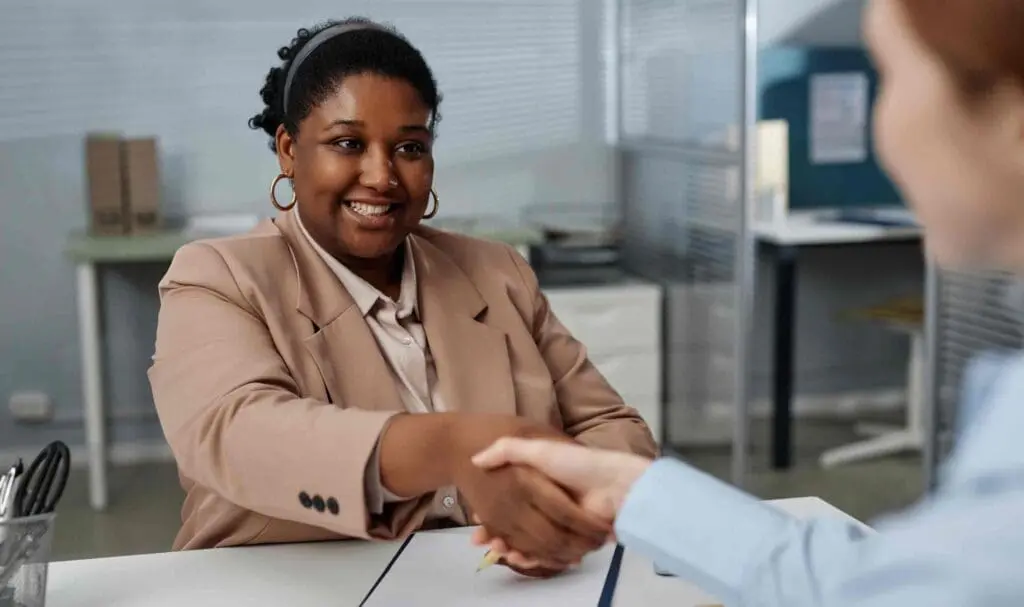 Une femme vêtue d'un blazer beige sourit et serre la main d'une autre personne assise à un bureau, suggérant un entretien d'embauche ou une réunion d'affaires réussi.