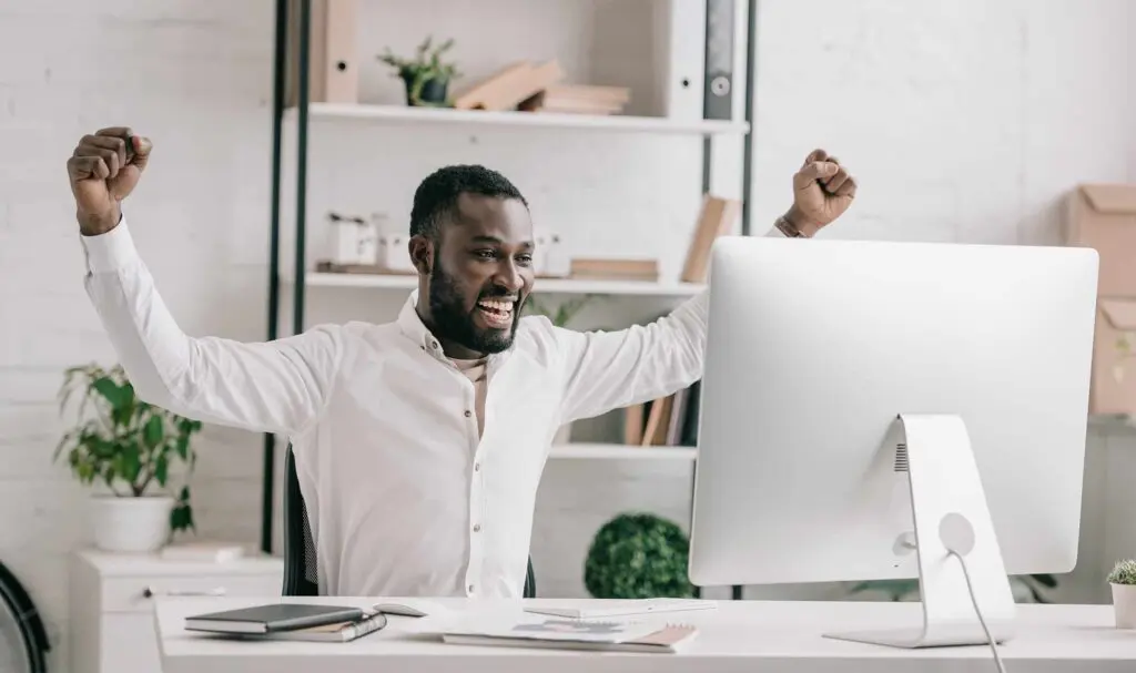 A man sitting at a desk in front of a computer, smiling and raising both arms in celebration, in a bright, modern office with shelves and plants in the background.