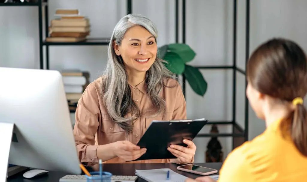 Une femme souriante aux longs cheveux gris tient un presse-papiers tout en parlant à une autre personne à travers un bureau dans un bureau avec des étagères et une plante en arrière-plan.