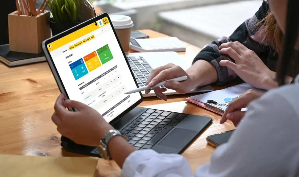 Deux personnes travaillent à un bureau en bois, une tablette posée sur un support pour clavier. L'une pointe un tableau de bord coloré sur l'écran avec un stylet, tandis que l'autre fait des gestes à proximité. Un carnet, une calculatrice et une tasse à café sont également posés sur le bureau.