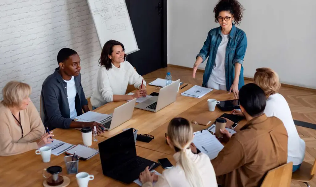 Un groupe de six personnes est assis autour d'une table avec des ordinateurs portables et des papiers, tandis qu'une personne se tient debout et fait des gestes, menant une discussion dans un cadre de bureau lumineux et moderne.