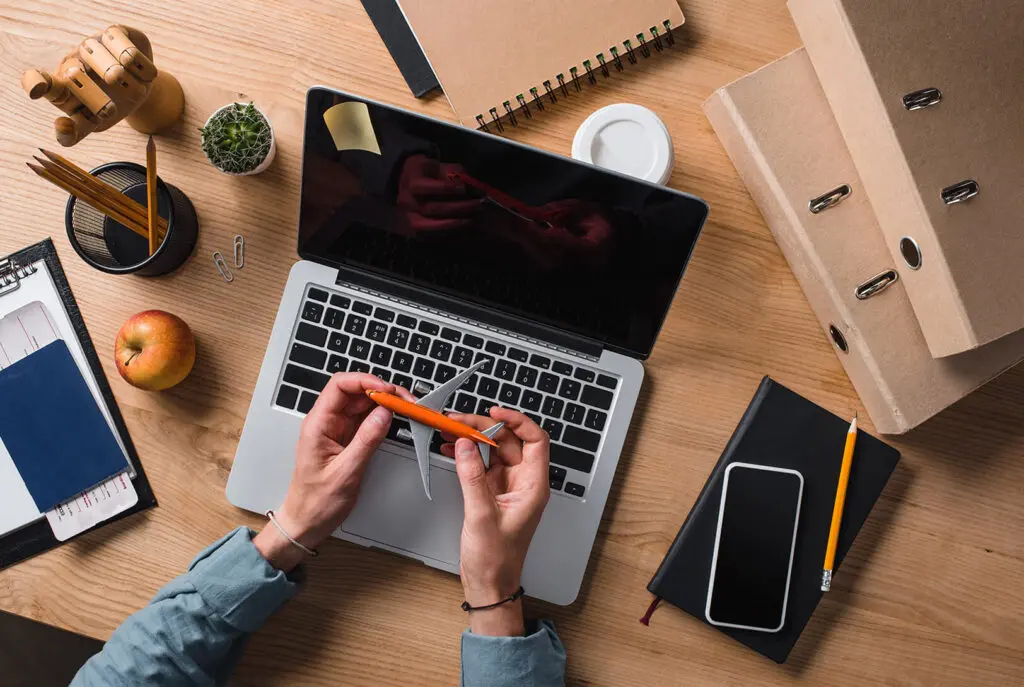 Vue de dessus d'une personne assise à un bureau avec un ordinateur portable, un stylo orange à la main. Le bureau contient des dossiers, des cahiers, un smartphone, une pomme, une tasse à café, une plante et des fournitures de bureau.