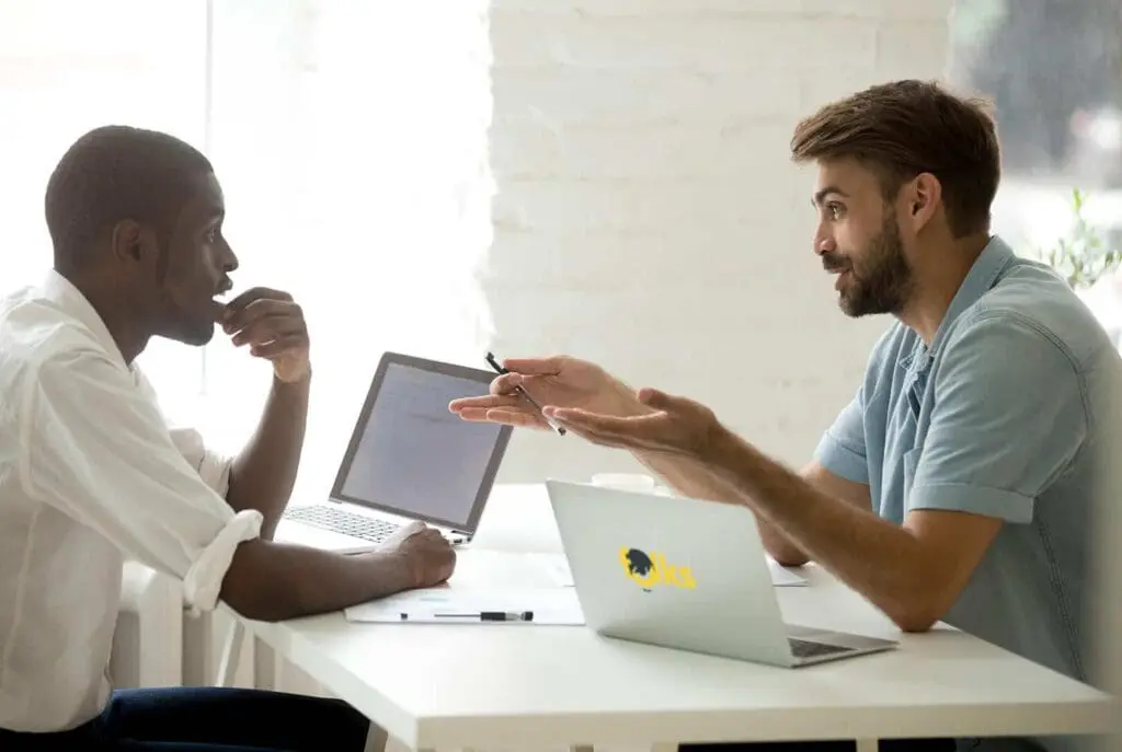 Deux hommes sont assis face à face à un bureau avec des ordinateurs portables, en pleine conversation. L'un fait des gestes avec un stylo, tandis que l'autre écoute pensivement, la main sur le menton.