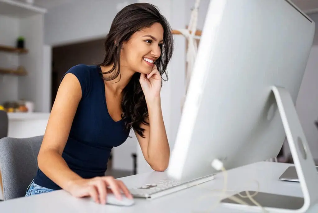 Une femme aux longs cheveux noirs est assise à un bureau, souriante devant un ordinateur. Elle porte une chemise bleu marine et semble travailler dans un bureau à domicile lumineux et moderne.