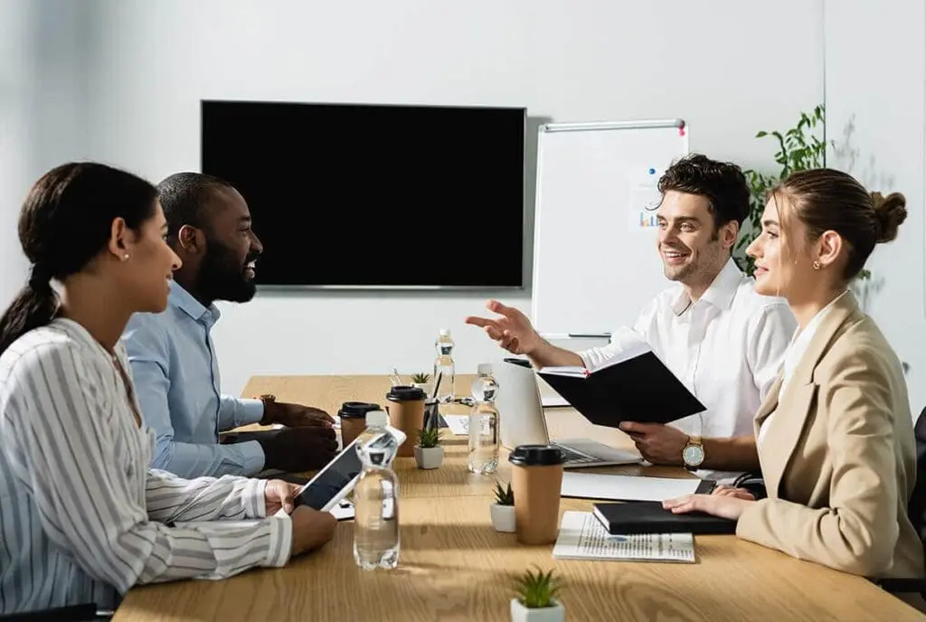 Quatre professionnels sont assis autour d'une table de conférence et discutent. Ils sourient, tenant des cahiers et des tablettes. Des bouteilles d'eau, des tasses à café et des plantes sont disposées sur la table, avec un écran de télévision vierge et un tableau de conférence en arrière-plan.