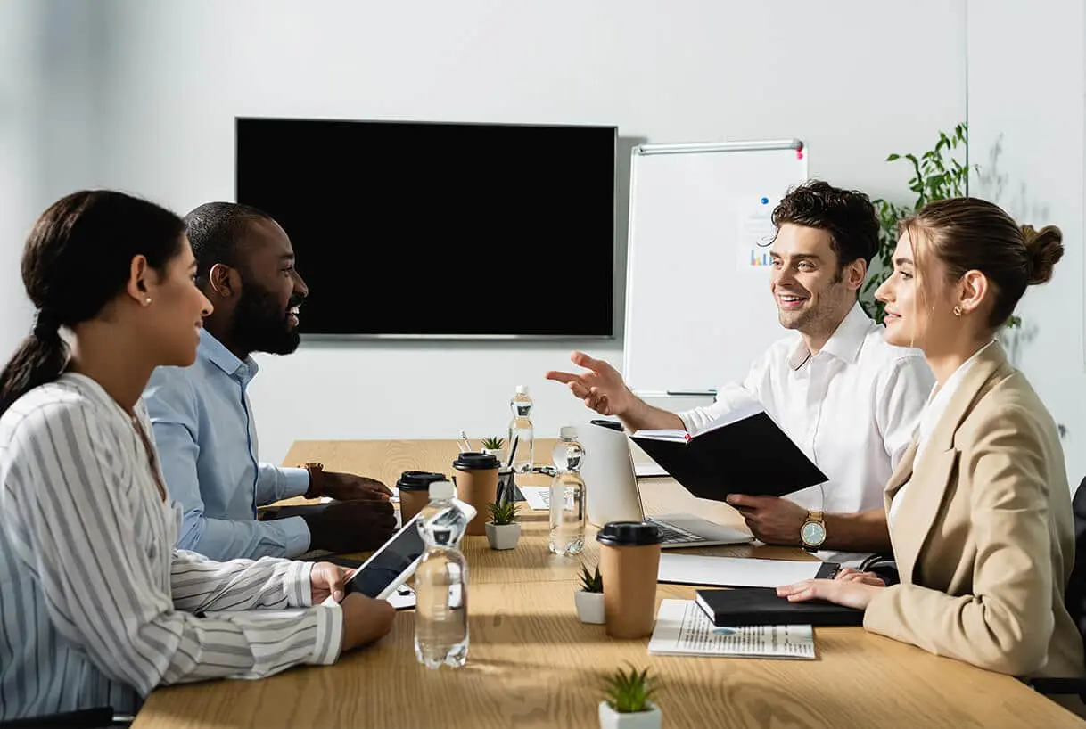 Quatre professionnels sont assis autour d'une table de conférence et discutent. Ils sourient, tenant des cahiers et des tablettes. Des bouteilles d'eau, des tasses à café et des plantes sont disposées sur la table, avec un écran de télévision vierge et un tableau de conférence en arrière-plan.