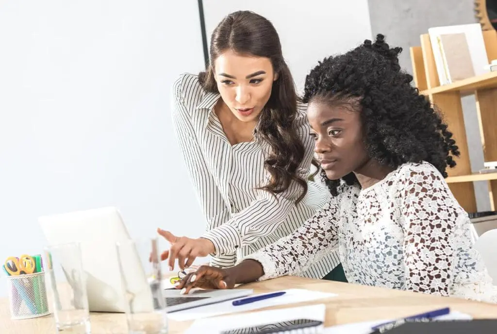 Deux femmes travaillent ensemble à un bureau, les yeux rivés sur l'écran d'un ordinateur portable. L'une est assise et tape du texte, tandis que l'autre se tient à côté d'elle, pointant l'écran du doigt et expliquant quelque chose. Des fournitures de bureau sont visibles sur le bureau.