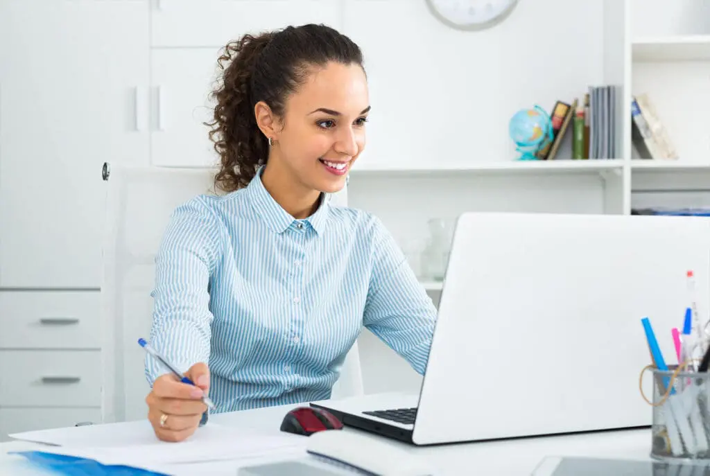 Une femme assise à un bureau, souriante devant son ordinateur portable, tient un stylo dans une main, tandis que des papiers et des fournitures de bureau sont posés sur le bureau. Des étagères et des livres sont visibles en arrière-plan.