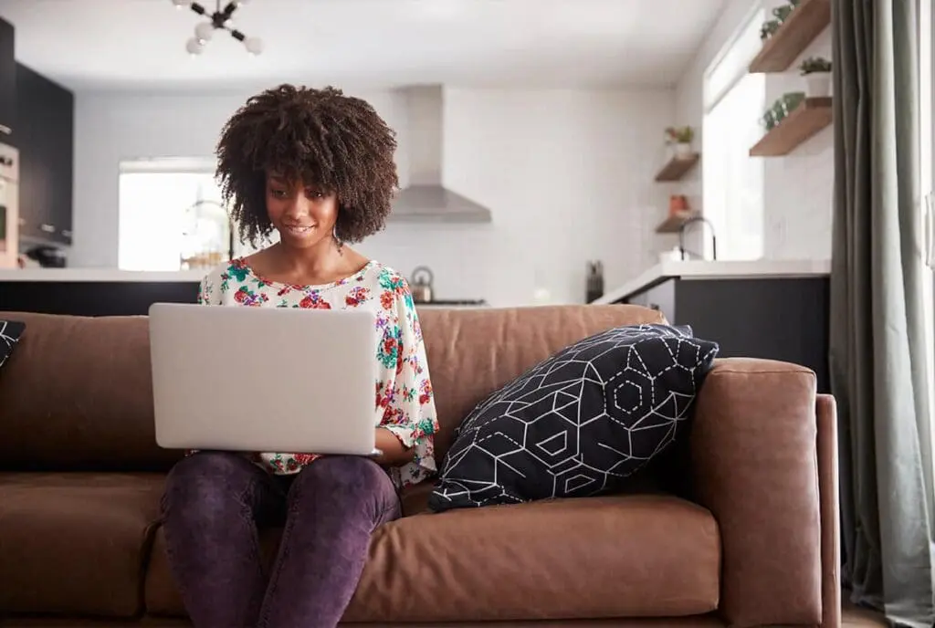 Une femme est assise sur un canapé marron dans un salon moderne, souriante, son ordinateur portable posé sur ses genoux. Un coussin à motifs est posé à côté d'elle et une cuisine est visible en arrière-plan.