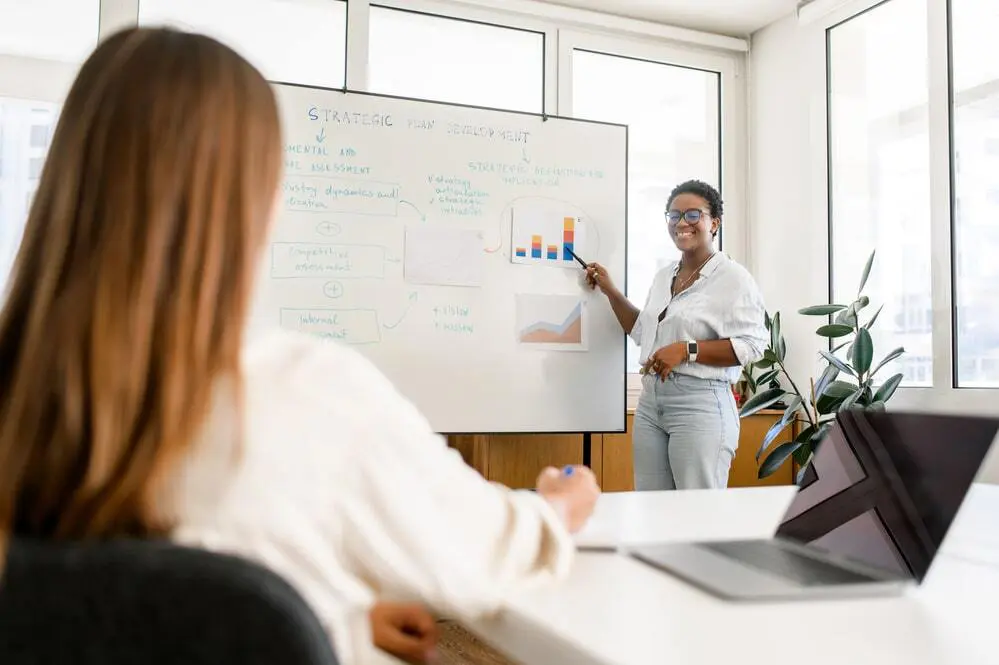 A woman stands by a whiteboard presenting charts and graphs to a colleague seated at a desk with a laptop in a bright office. The presenter is smiling and holding a marker.