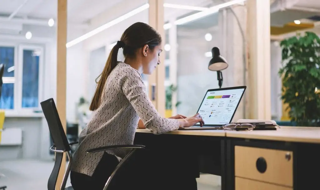 Une femme coiffée d'une queue de cheval est assise à un bureau dans un bureau moderne, travaillant sur un ordinateur portable. Une lampe de bureau, un téléphone et un carnet sont à proximité, tandis que de grandes fenêtres et des plantes sont visibles en arrière-plan.