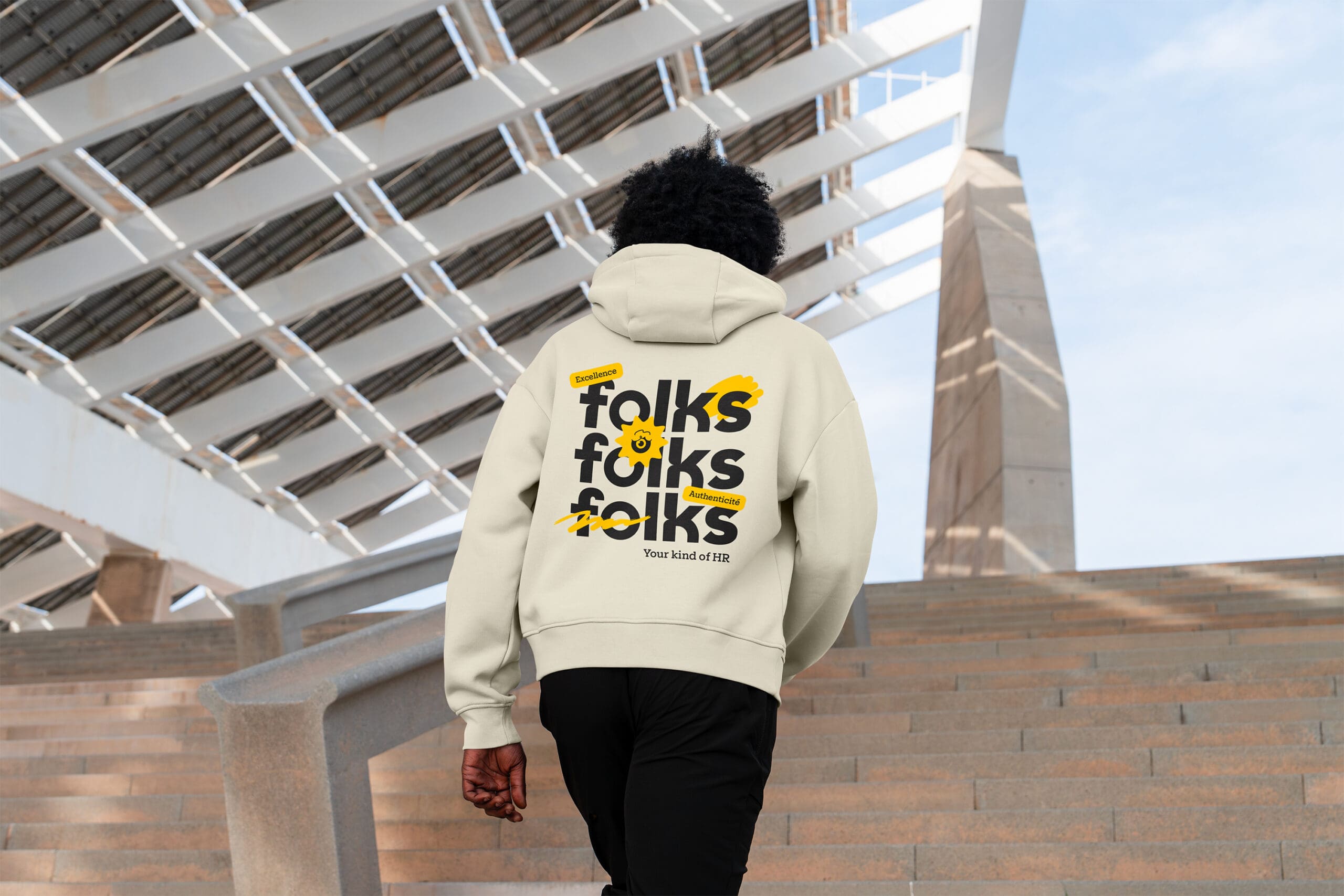 Une personne portant un sweat à capuche de couleur claire avec le mot « folks » répété trois fois dans le dos, montant des marches extérieures sous une structure géométrique moderne.
