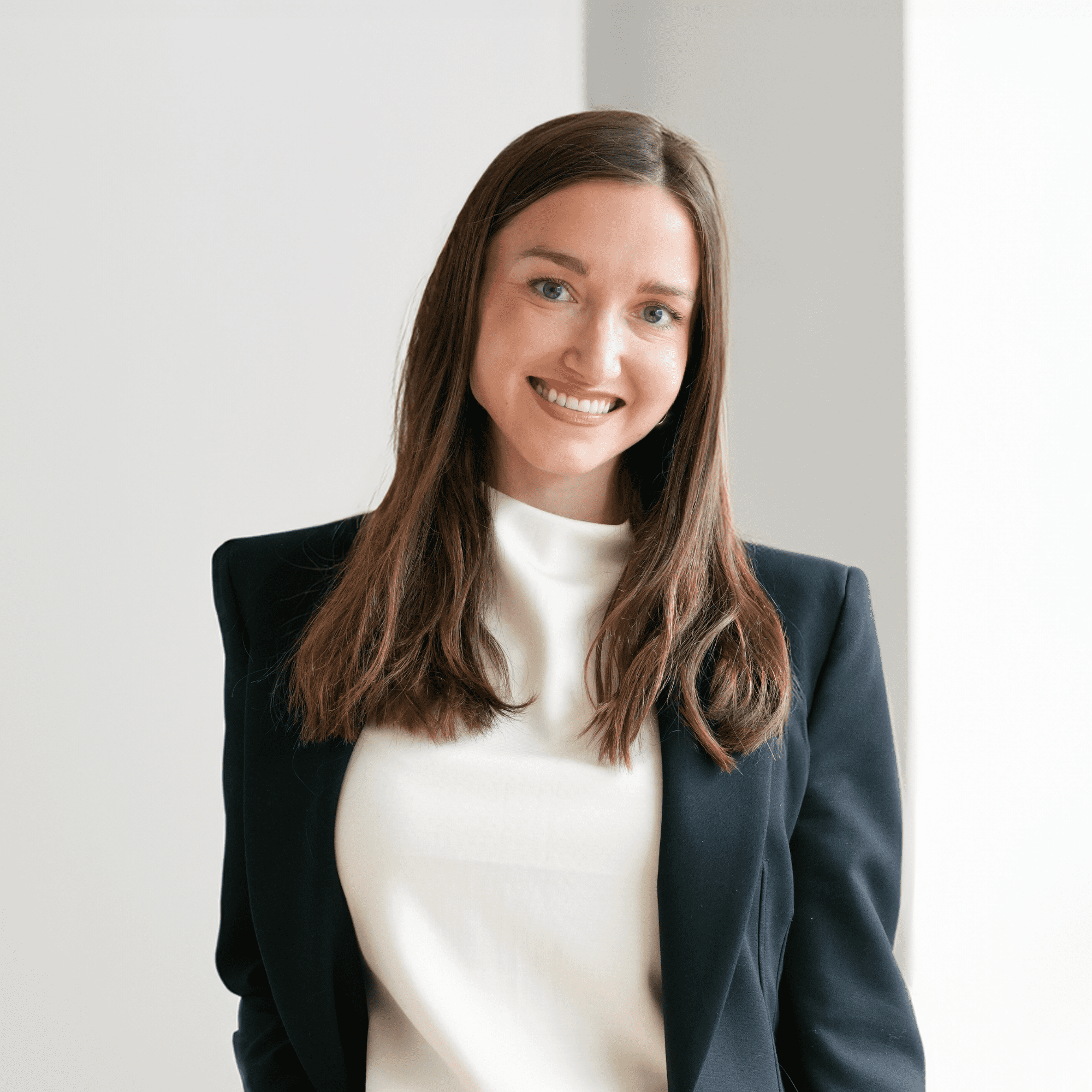 Une jeune femme aux longs cheveux bruns, portant un blazer noir sur un haut blanc, souriante et debout dans un décor intérieur lumineux et minimaliste.