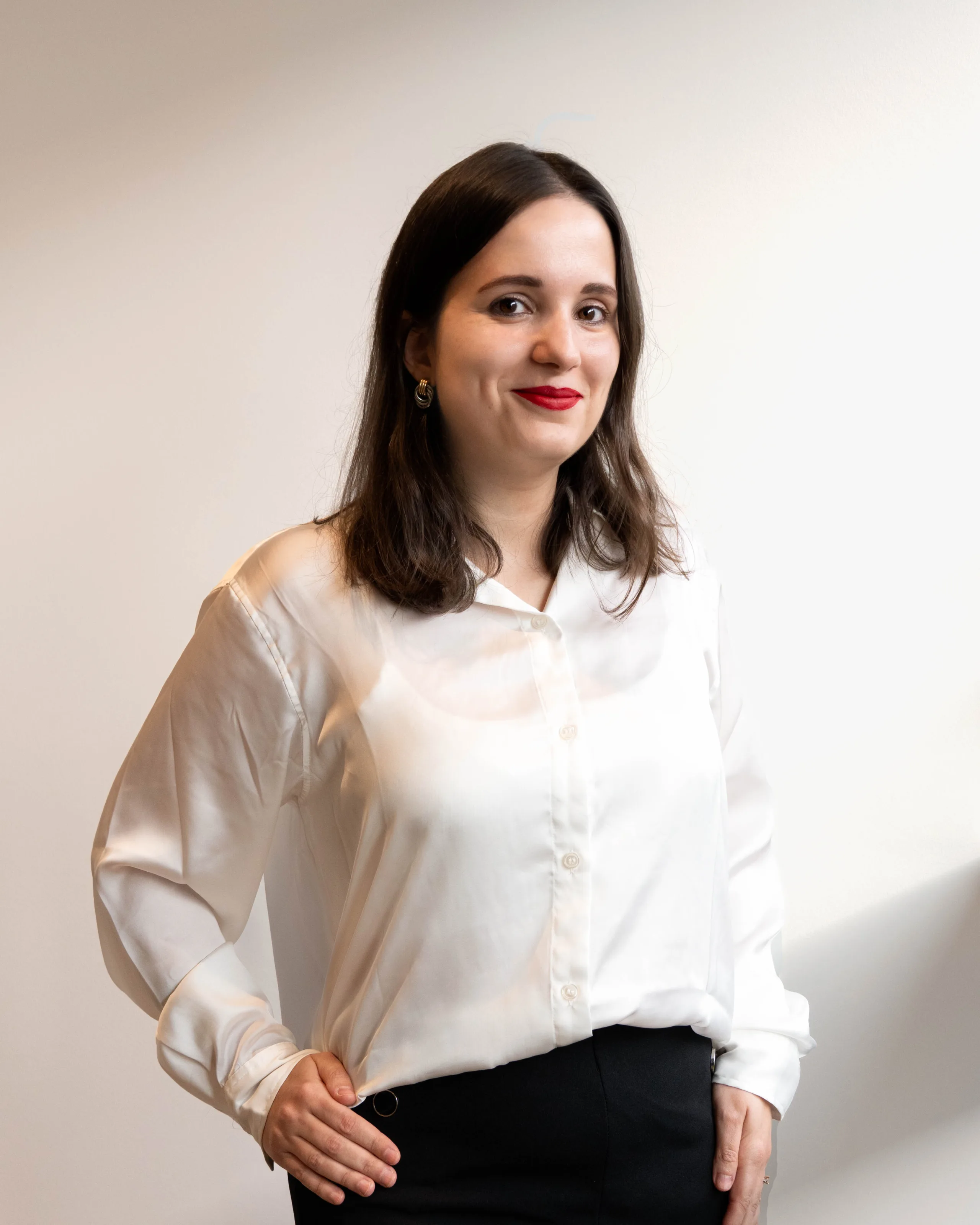 A woman with long dark hair, wearing a white button-up shirt and black pants, stands against a plain light background, smiling slightly with one hand on her hip.