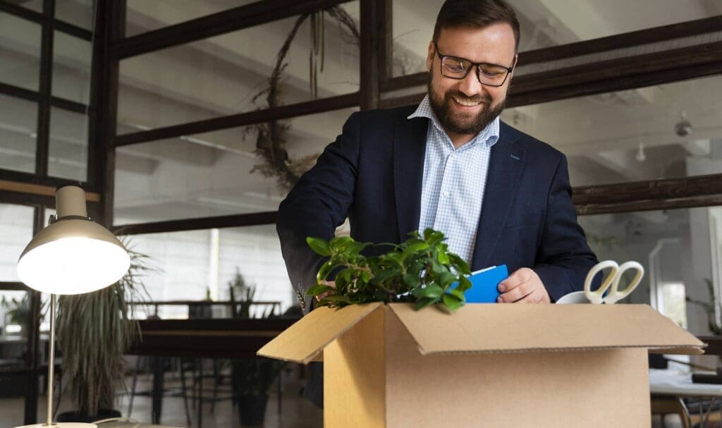 Un homme souriant en costume déballe une boîte en carton contenant des fournitures de bureau et une plante en pot sur un bureau dans un espace de travail moderne.