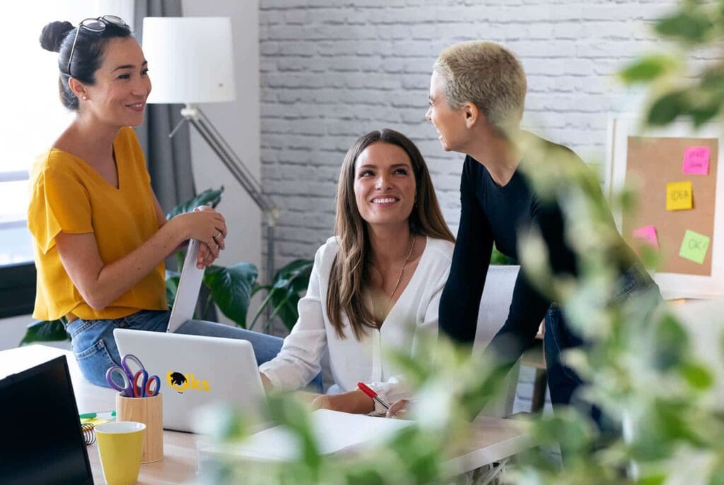Trois femmes en tenue de bureau décontractée sourient et discutent autour d'un bureau avec un ordinateur portable, des papiers et du matériel de bureau. L'une est assise, les deux autres se tiennent à proximité. Espace de bureau lumineux et moderne avec des plantes au premier plan.
