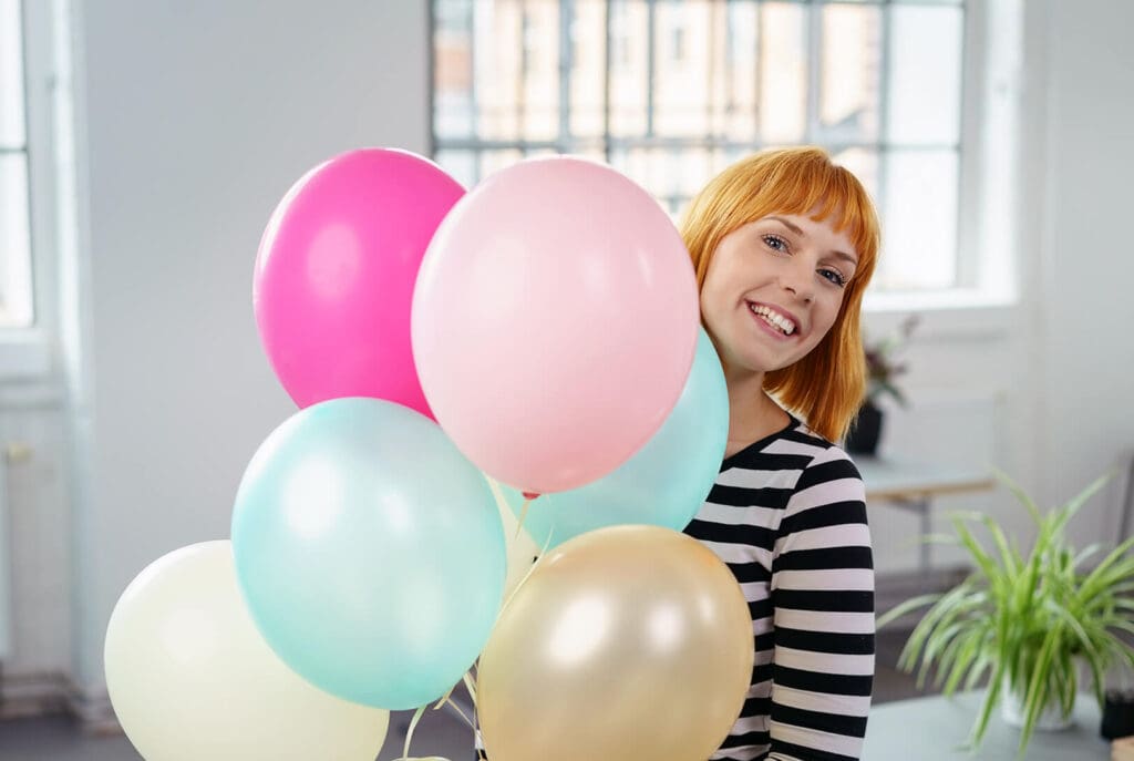 Une personne souriante aux cheveux roux courts, vêtue d'une chemise rayée noire et blanche, tient un bouquet de ballons colorés à l'intérieur. De grandes fenêtres et une plante verte sont visibles en arrière-plan.