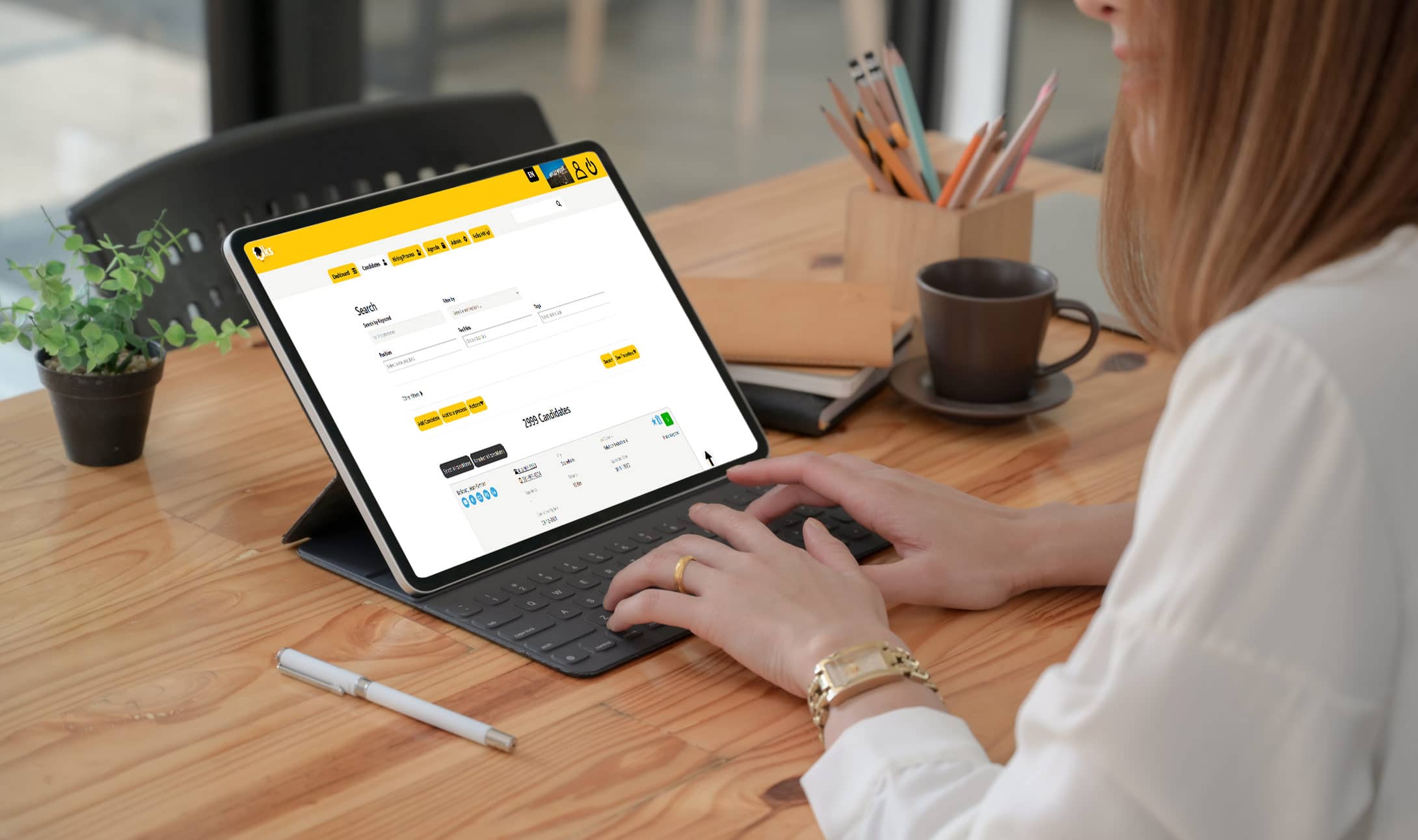 A person types on a tablet with a keyboard at a wooden desk, viewing a website with a yellow and white interface. Nearby are a notebook, a cup of coffee, and a pen.