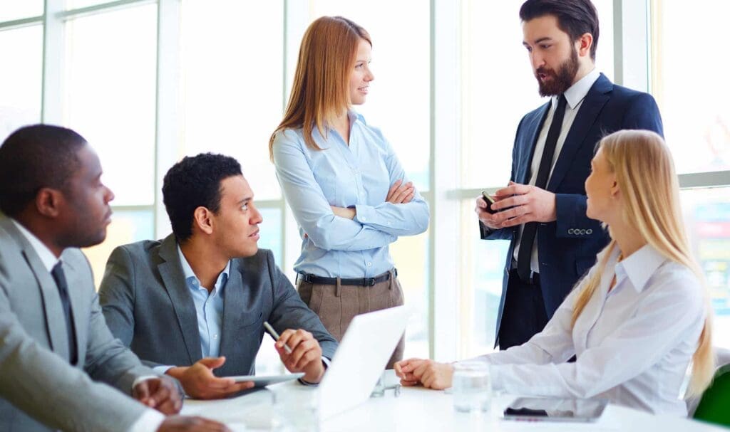 Cinq professionnels en tenue de soirée discutent autour d'une table dans un bureau lumineux. Un homme parle debout, tandis que les autres sont assis et écoutent attentivement. Ordinateurs portables et documents sont visibles.