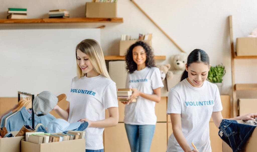 Trois femmes portant des t-shirts « BÉNÉVOLES » trient et organisent des dons, tels que des vêtements et des livres, dans une pièce lumineuse remplie de cartons et d'étagères. Souriantes et concentrées sur leur tâche, elles sont à l'écoute.