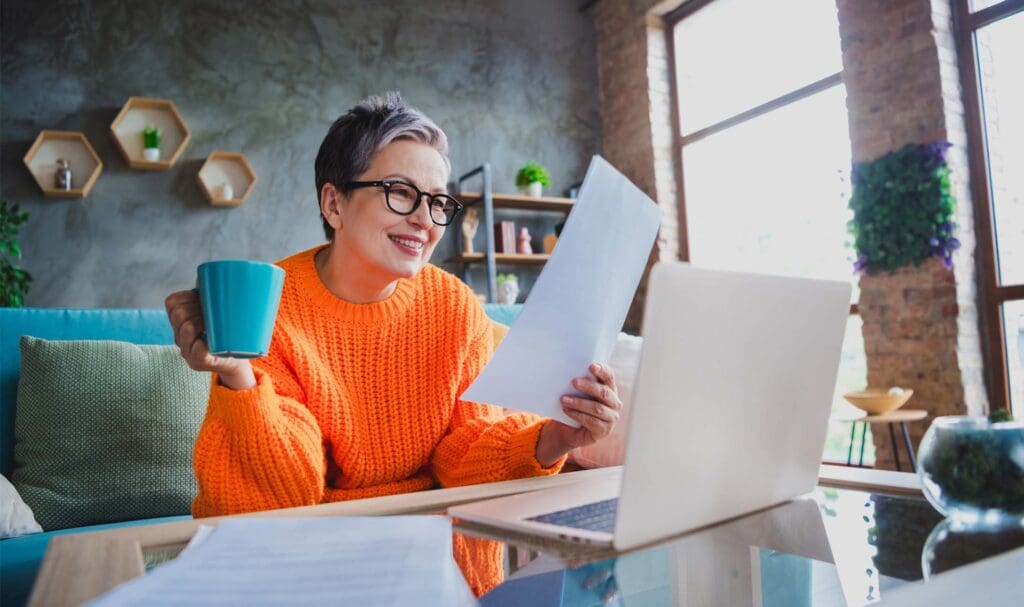 An older woman wearing glasses and an orange sweater sits on a couch, smiling while holding a blue mug and reading a document in front of an open laptop in a bright, modern living room.