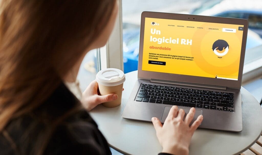 Une personne tenant une tasse de café en papier utilise un ordinateur portable affichant un site Web jaune vif en français sur les logiciels RH abordables, assise à une petite table ronde près d'une fenêtre.