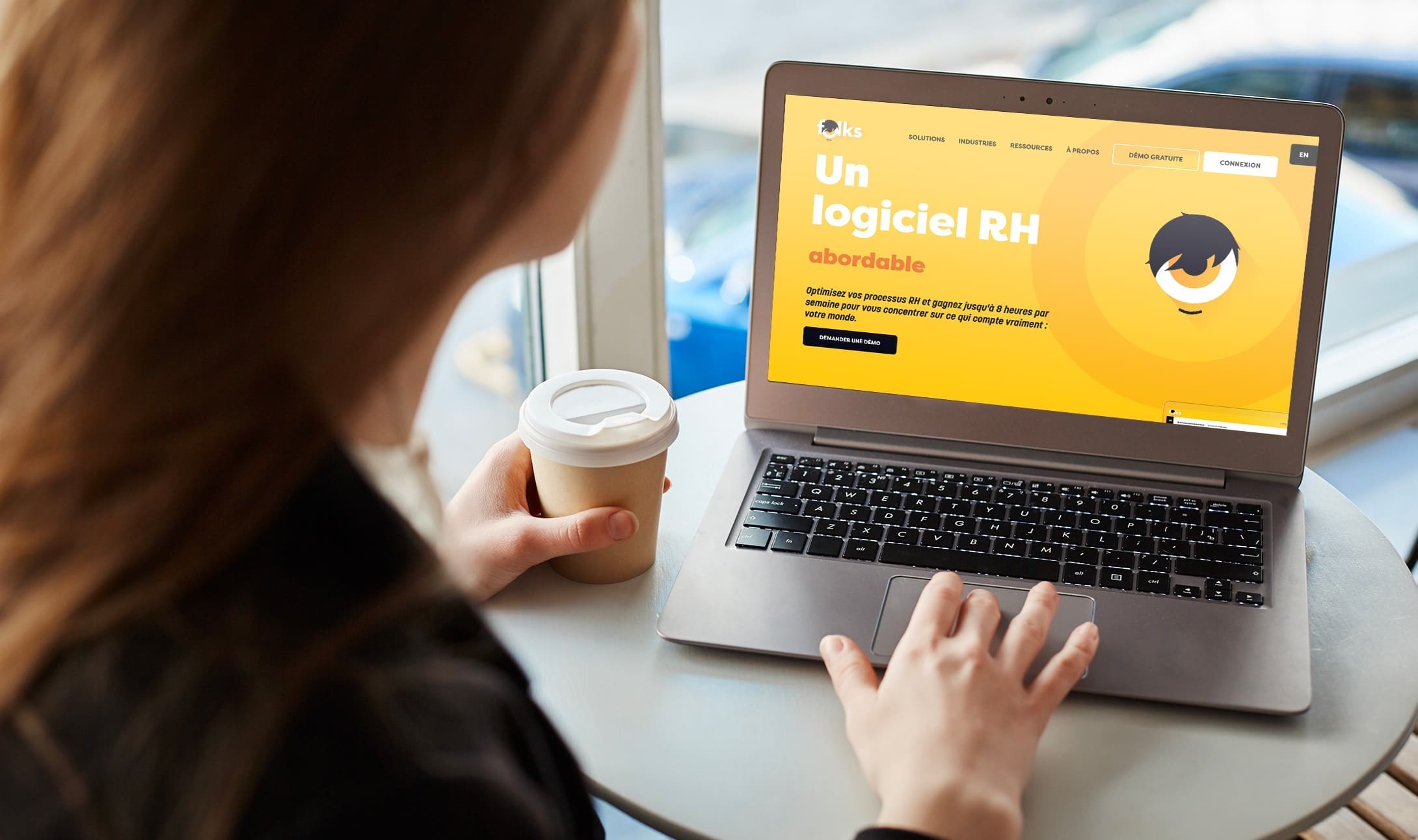 Une personne tenant une tasse de café en papier utilise un ordinateur portable affichant un site Web jaune vif en français sur les logiciels RH abordables, assise à une petite table ronde près d'une fenêtre.