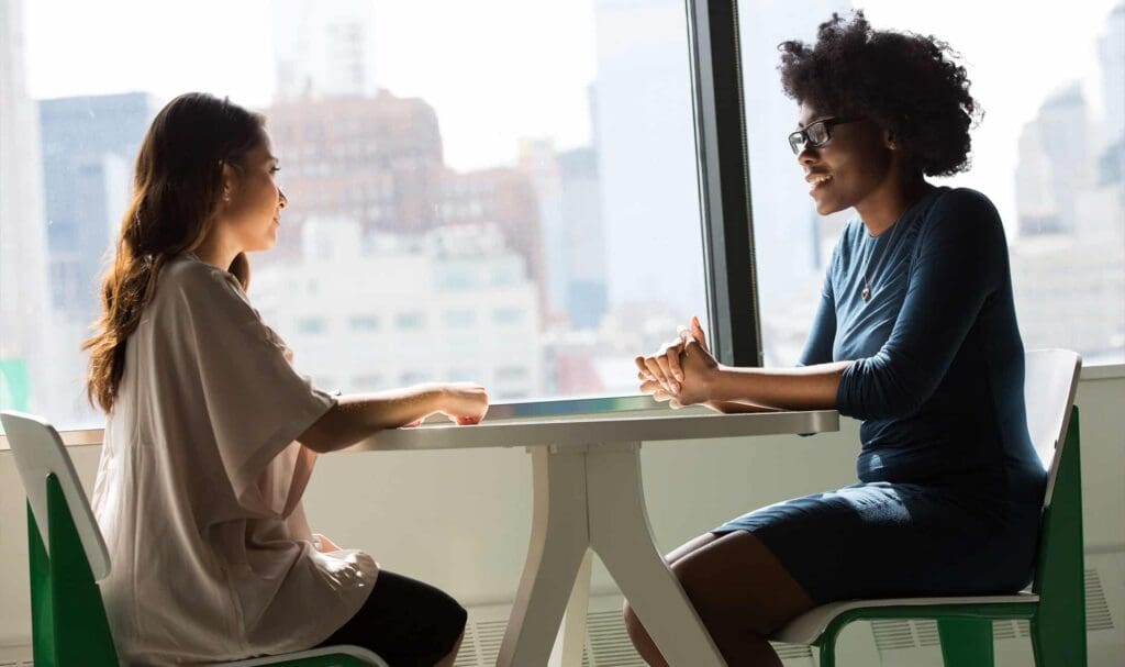 Two women sit across from each other at a table by a large window, engaged in conversation in a bright, modern office setting with city buildings visible in the background.
