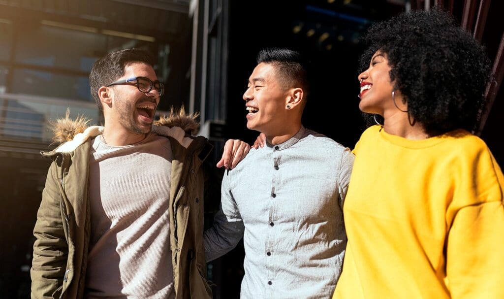 Trois amis se promènent dehors au soleil, souriant et riant ensemble. L'homme à gauche porte des lunettes et un manteau, celui du milieu une chemise claire et la femme à droite un pull jaune vif.