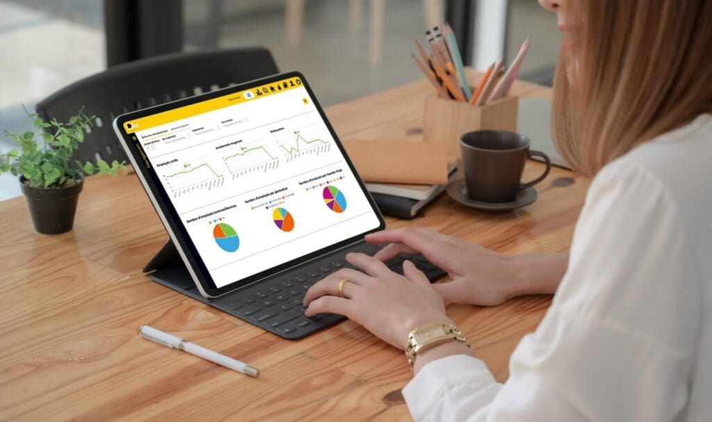 Une personne travaille sur une tablette avec clavier, affichant des tableaux et des graphiques. La tablette est posée sur un bureau en bois, avec un stylo, une tasse à café, une plante en pot et un porte-stylo à proximité.