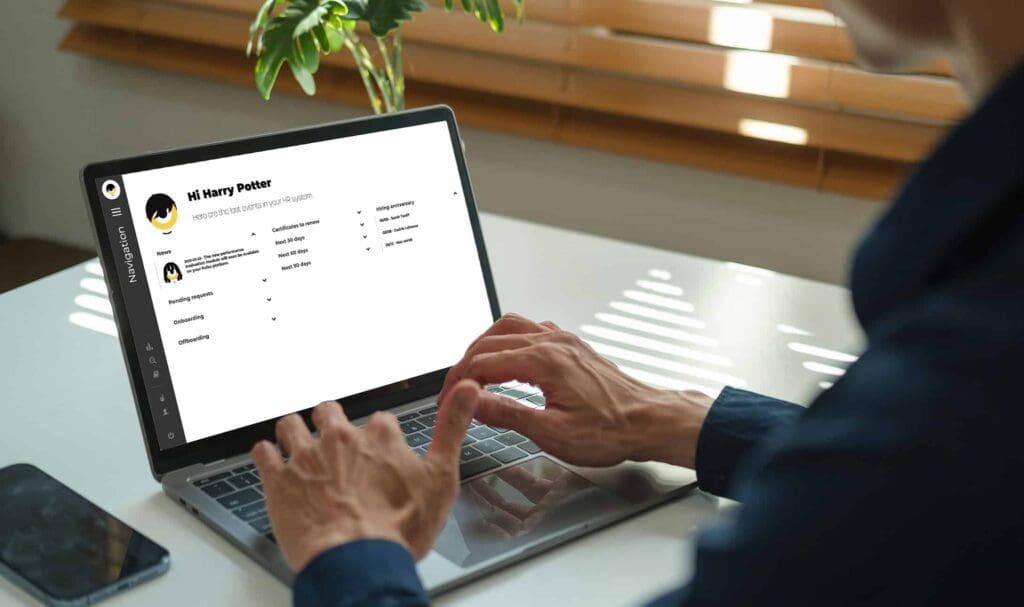 A person wearing a dark shirt types on a laptop at a white desk. The laptop screen displays a profile page with the name