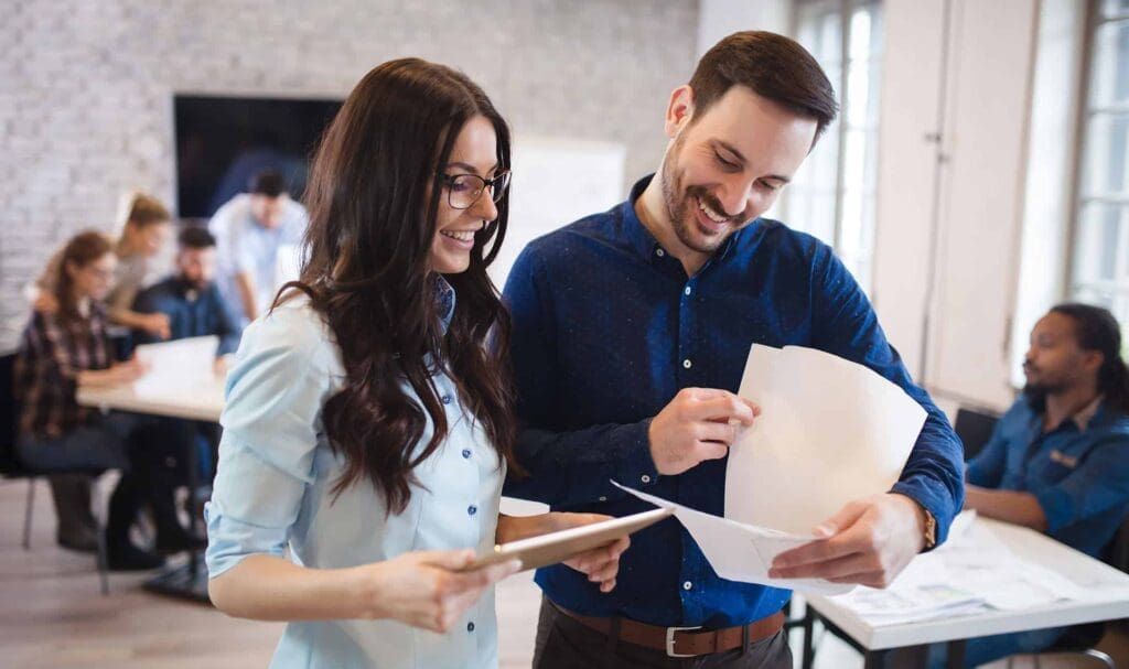Deux collègues se tiennent ensemble dans un bureau, souriant et examinant des documents. D'autres collègues sont assis et travaillent en arrière-plan, créant une ambiance de travail collaborative et conviviale.