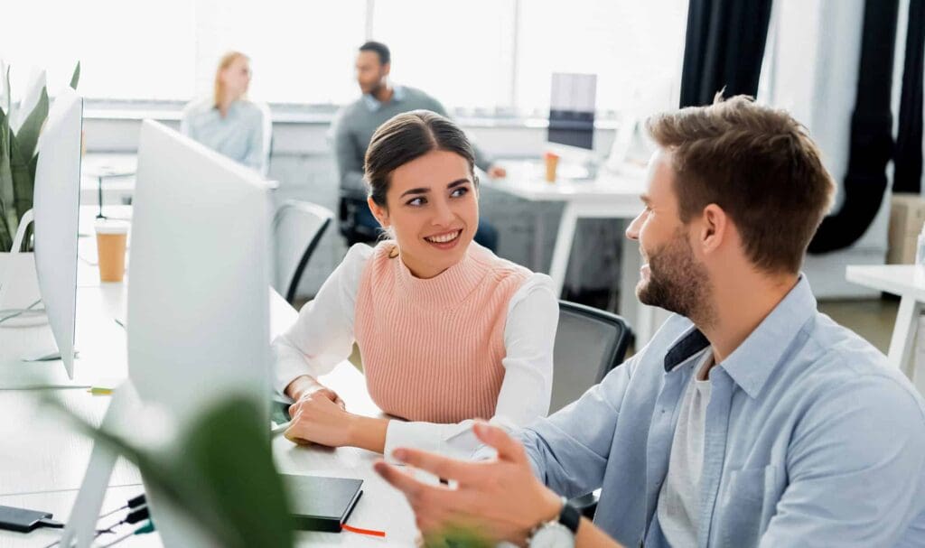 Two coworkers sit at a desk in a bright office, smiling and talking to each other in front of a computer monitor, with two more colleagues in the background.