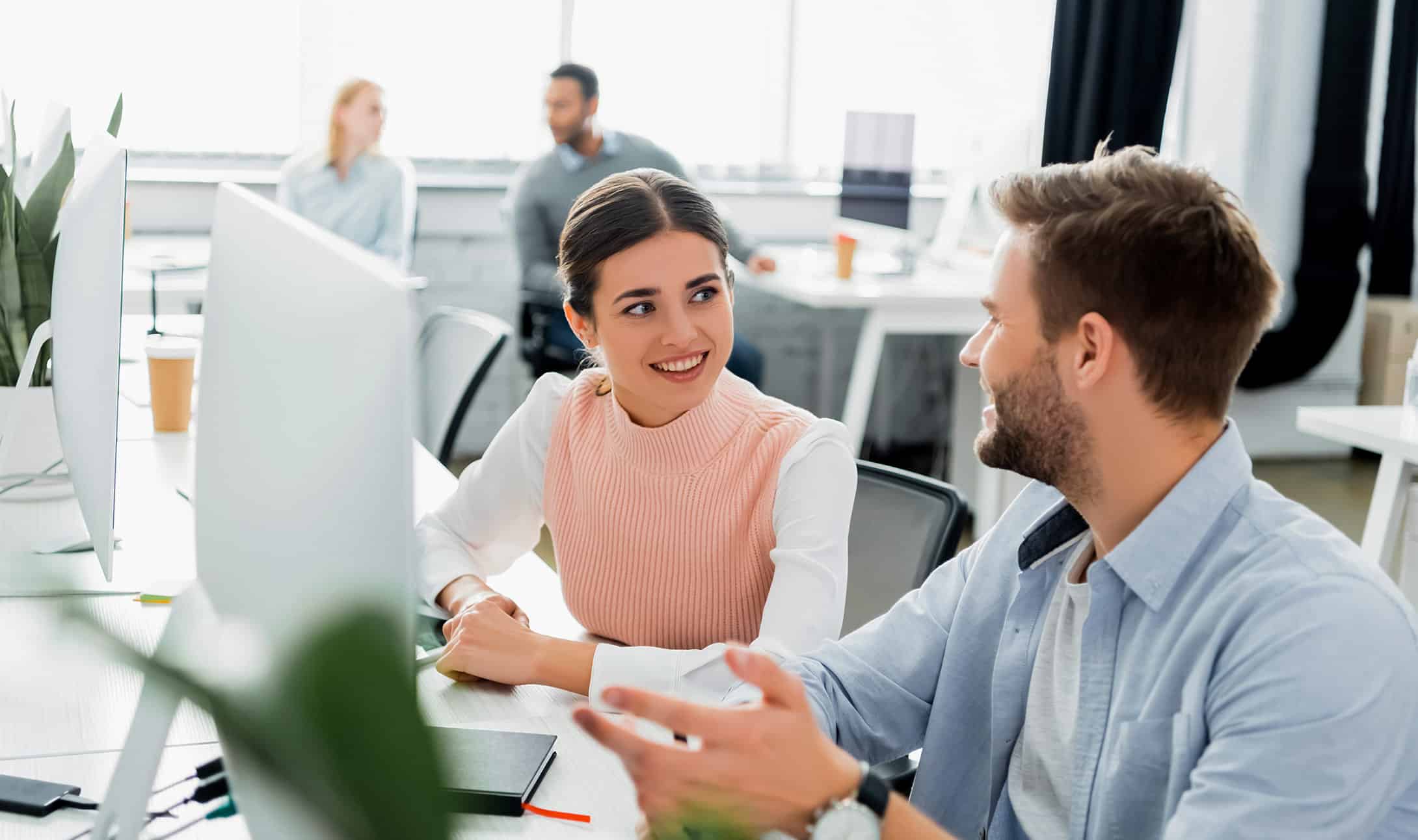 Two coworkers sit at a desk in a bright office, smiling and talking to each other in front of a computer monitor, with two more colleagues in the background.