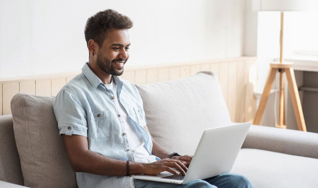 A smiling man sits on a beige sofa using a laptop, wearing a light denim shirt and blue jeans. Natural light enters from a window beside him, and a tripod lamp stands in the background.