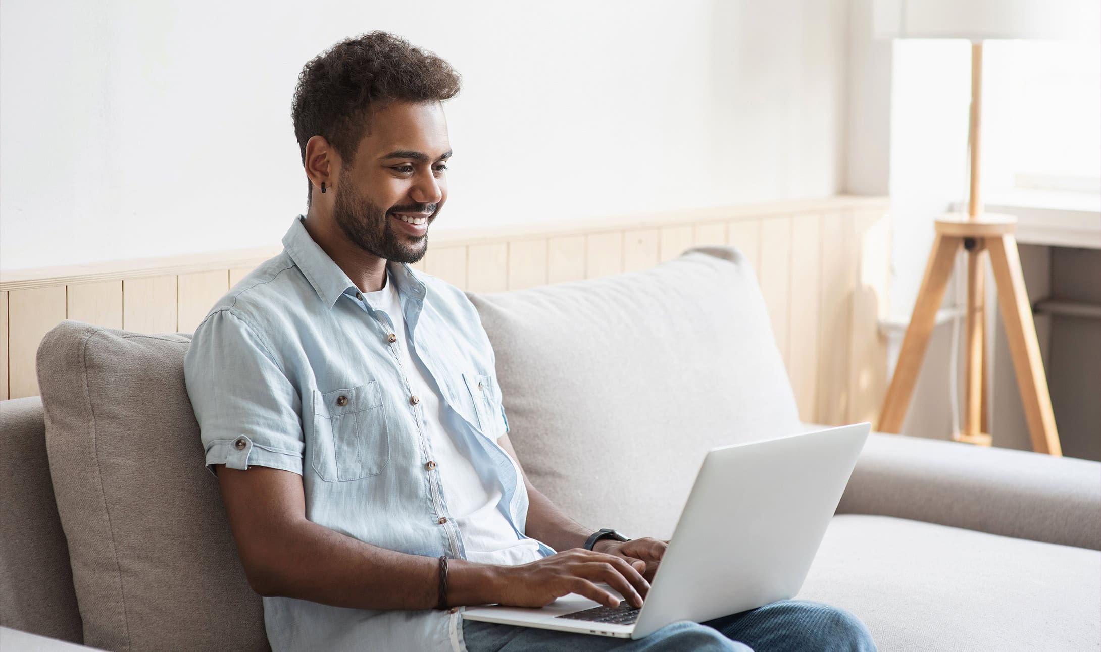 A smiling man sits on a beige sofa using a laptop, wearing a light denim shirt and blue jeans. Natural light enters from a window beside him, and a tripod lamp stands in the background.