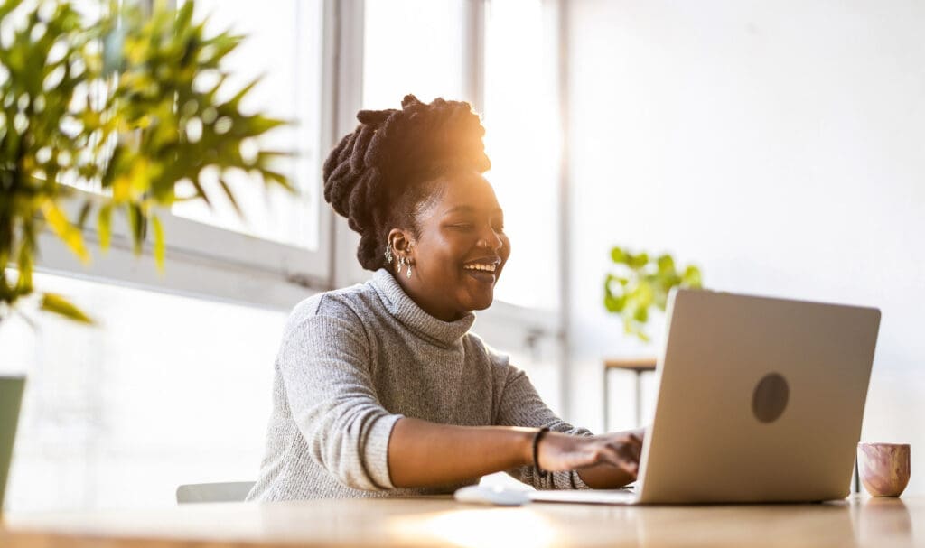 Une femme aux cheveux tressés sourit en travaillant sur son ordinateur portable, installée à un bureau lumineux et moderne. La lumière du soleil entre par de grandes fenêtres et des plantes en pot décorent l'espace.