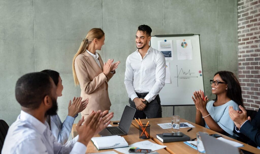Un groupe de collègues en tenue professionnelle est assis autour d'une table, applaudissant et souriant à un homme debout lors d'une réunion de bureau. Un tableau de conférence avec des graphiques est visible en arrière-plan.