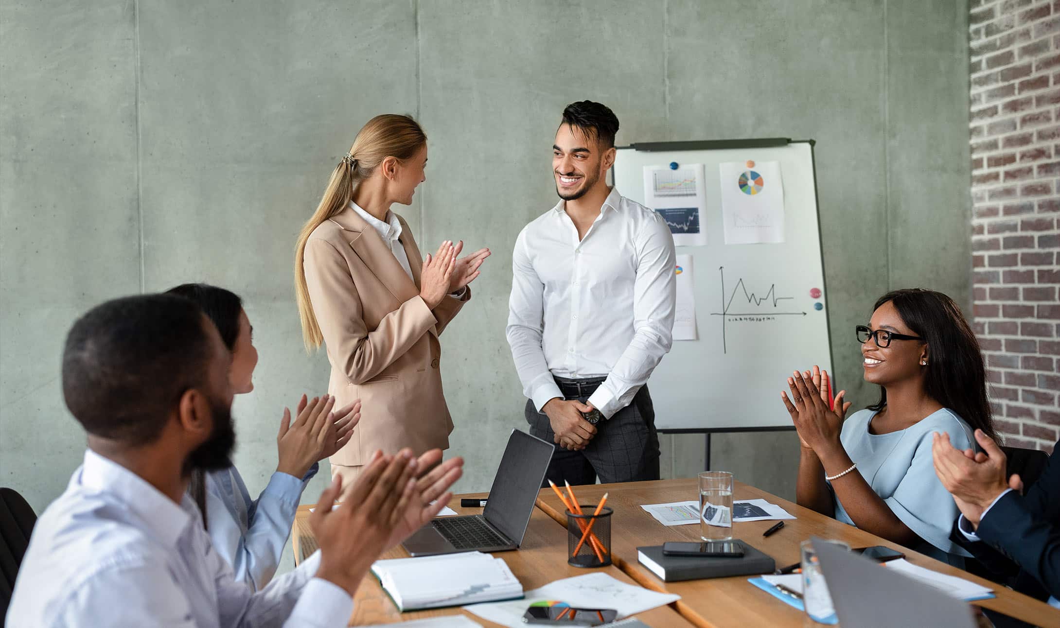 Un groupe de collègues en tenue professionnelle est assis autour d'une table, applaudissant et souriant à un homme debout lors d'une réunion de bureau. Un tableau de conférence avec des graphiques est visible en arrière-plan.