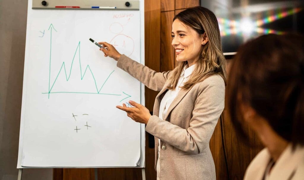Une femme vêtue d'un blazer beige se tient à côté d'un tableau à feuilles mobiles, souriant et pointant du doigt un graphique linéaire tout en faisant une présentation à une autre personne dans un bureau.
