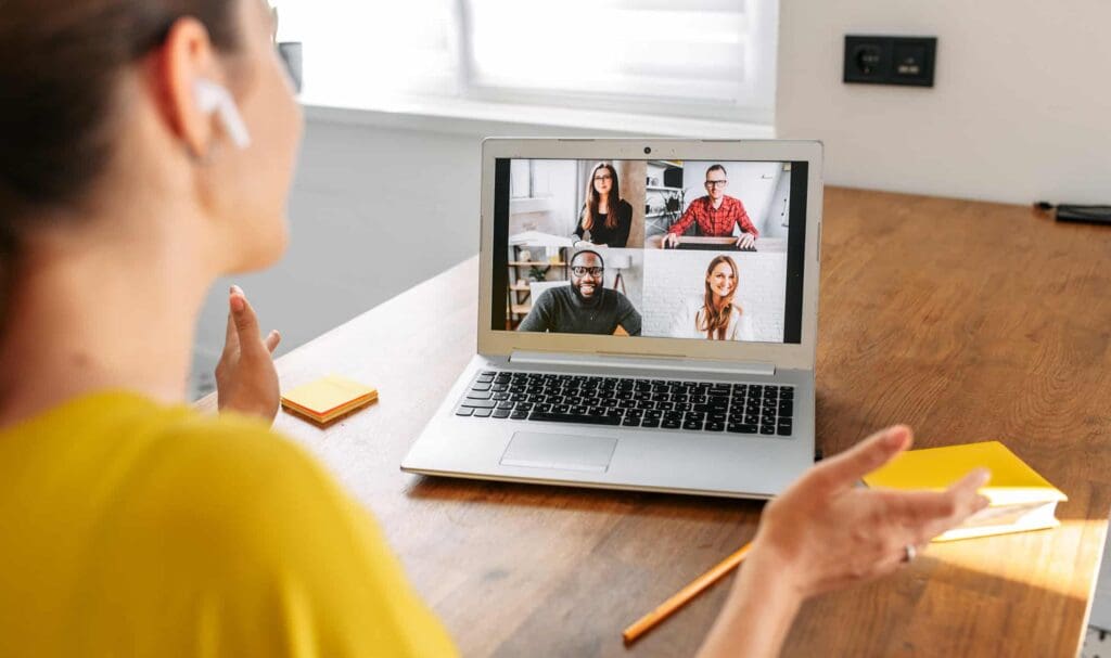 A woman wearing earbuds sits at a desk, participating in a video call on her laptop with four people visible on the screen. Sunlight comes through a window in the background.